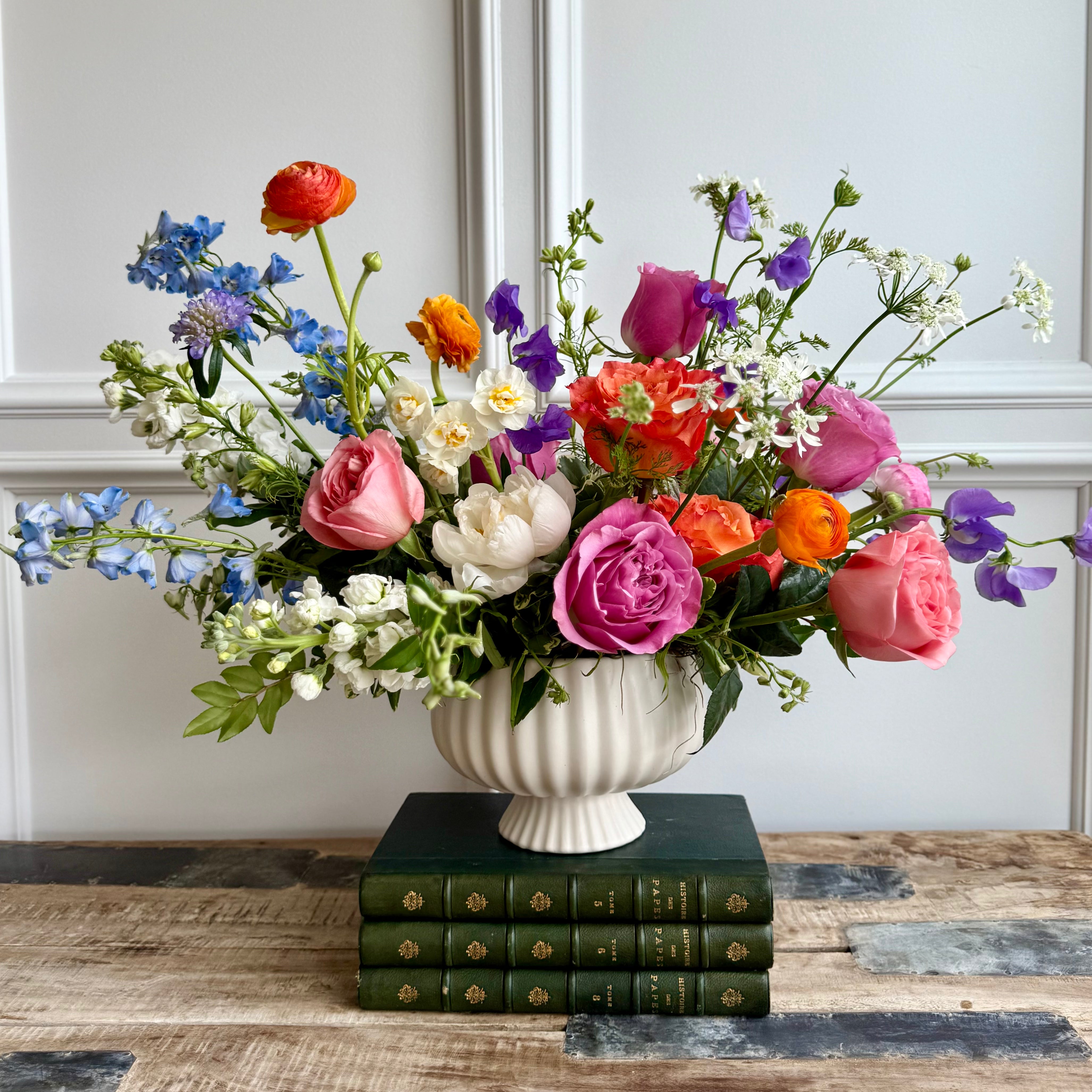 vibrant floral arrangement in a white ceramic vase featuring pink, orange, and purple blooms including roses, ranunculus, sweet peas, and peonies, with touches of white and blue flowers. The bouquet is displayed on stacked green vintage books atop a rustic wooden table, with a white panel wall in the background.