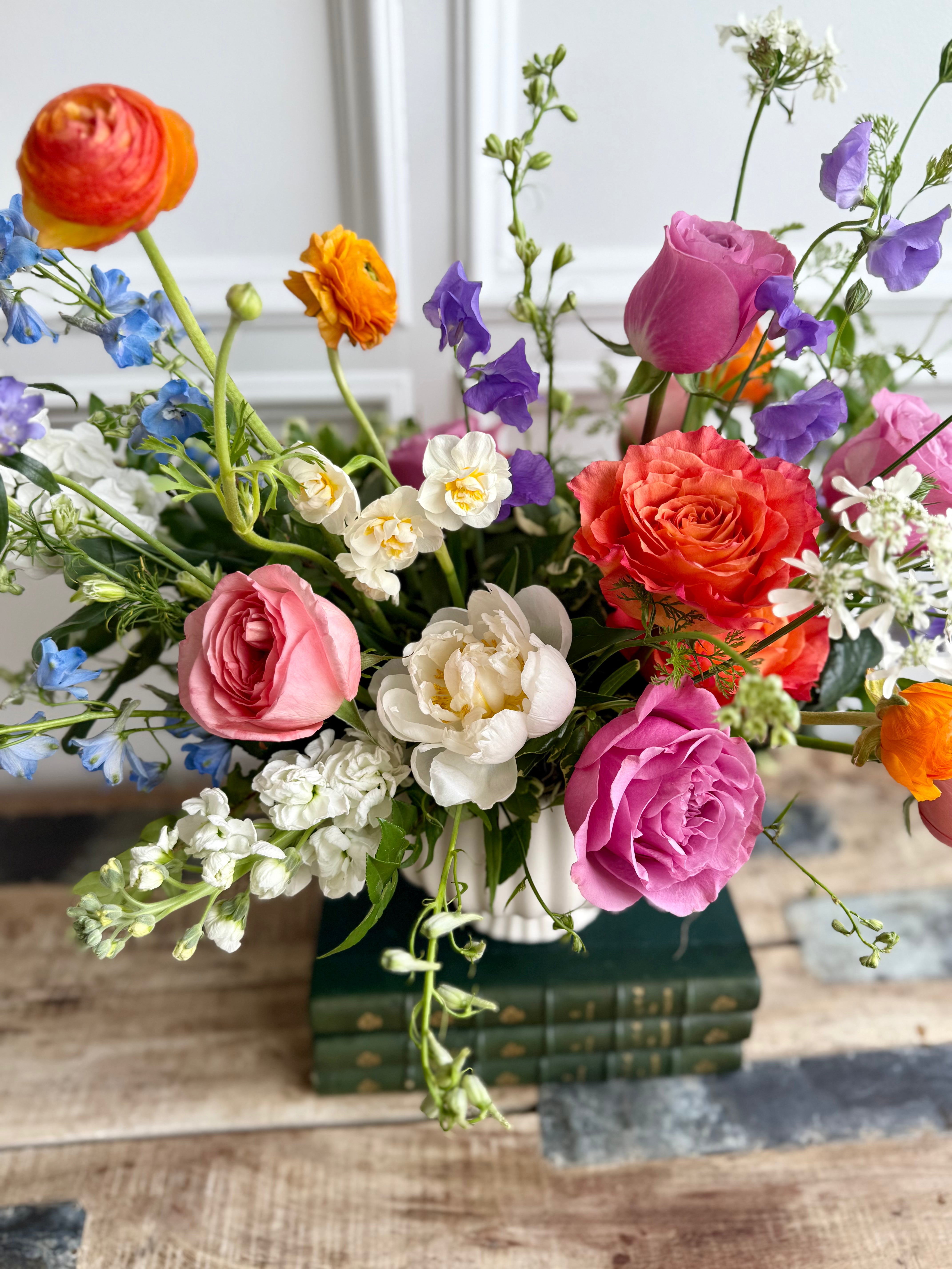 vibrant floral arrangement in a white ceramic vase featuring pink, orange, and purple blooms including roses, ranunculus, sweet peas, and peonies, with touches of white and blue flowers. The bouquet is displayed on stacked green vintage books atop a rustic wooden table, with a white panel wall in the background.