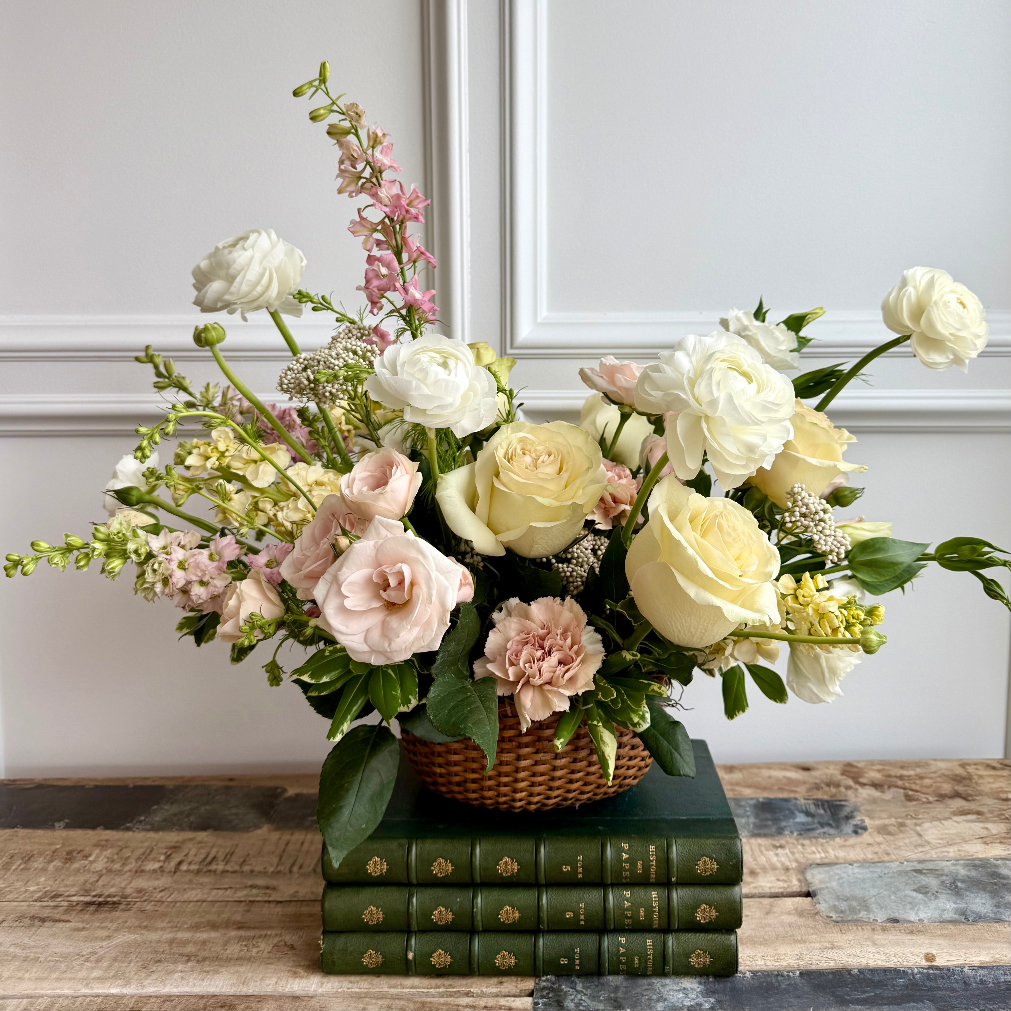 A soft and elegant floral arrangement in a woven basket, featuring cream and blush roses, white ranunculus, and delicate pink and ivory blooms. The bouquet is set on stacked green vintage books on a rustic wooden table, with a white panel wall in the background.
