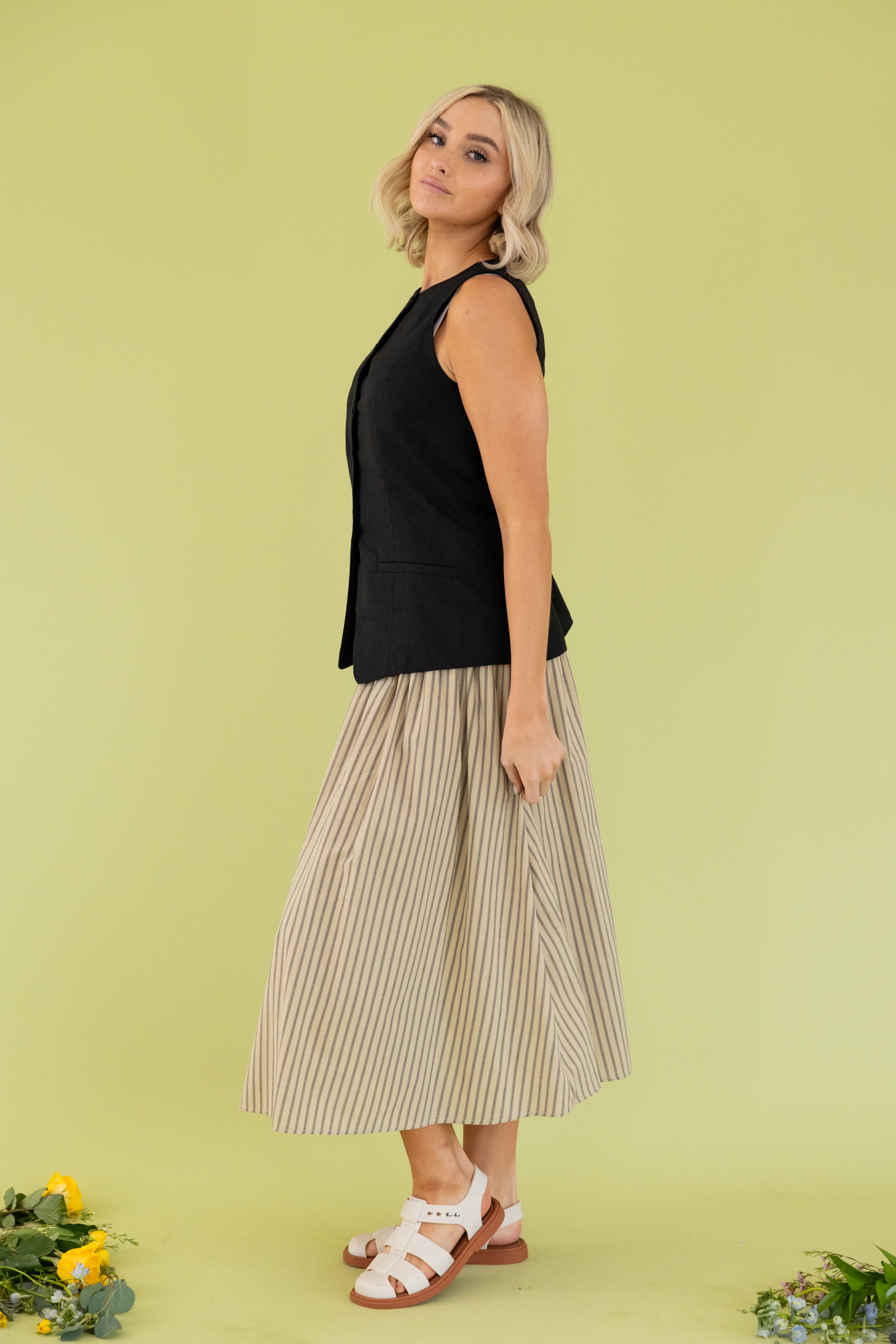 Model is wearing black vest with smocked tan striped midi skirt.