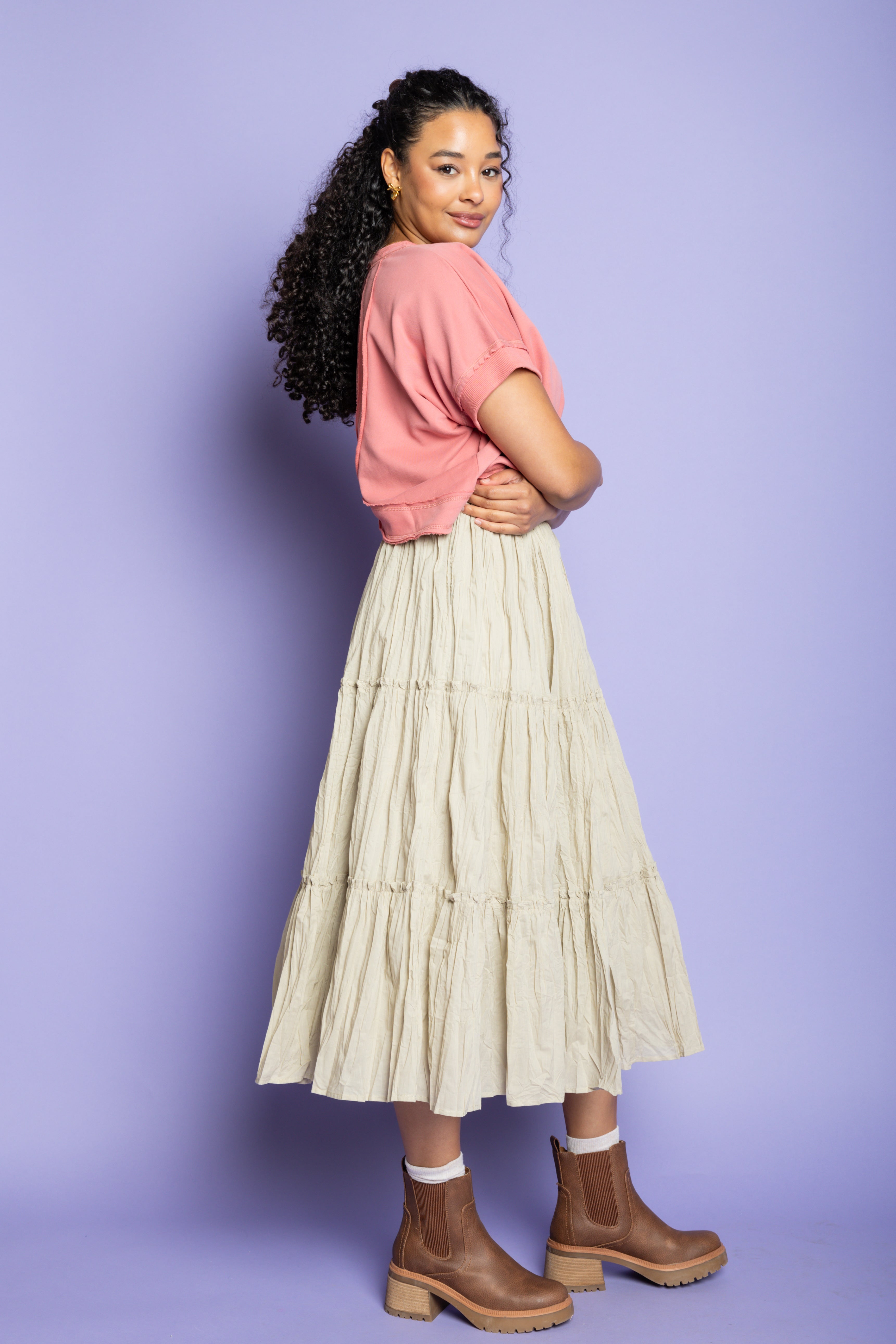 Model is wearing a pink t-shirt with tan ruffle skirt and brown boots.