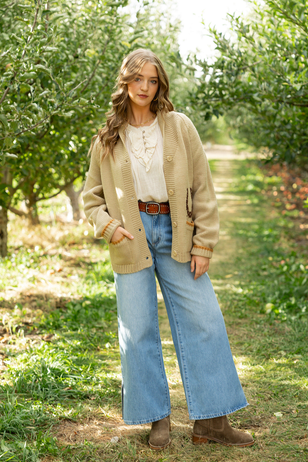 Woman standing in apple orchard. Wearing fall outfit and wide leg pants.