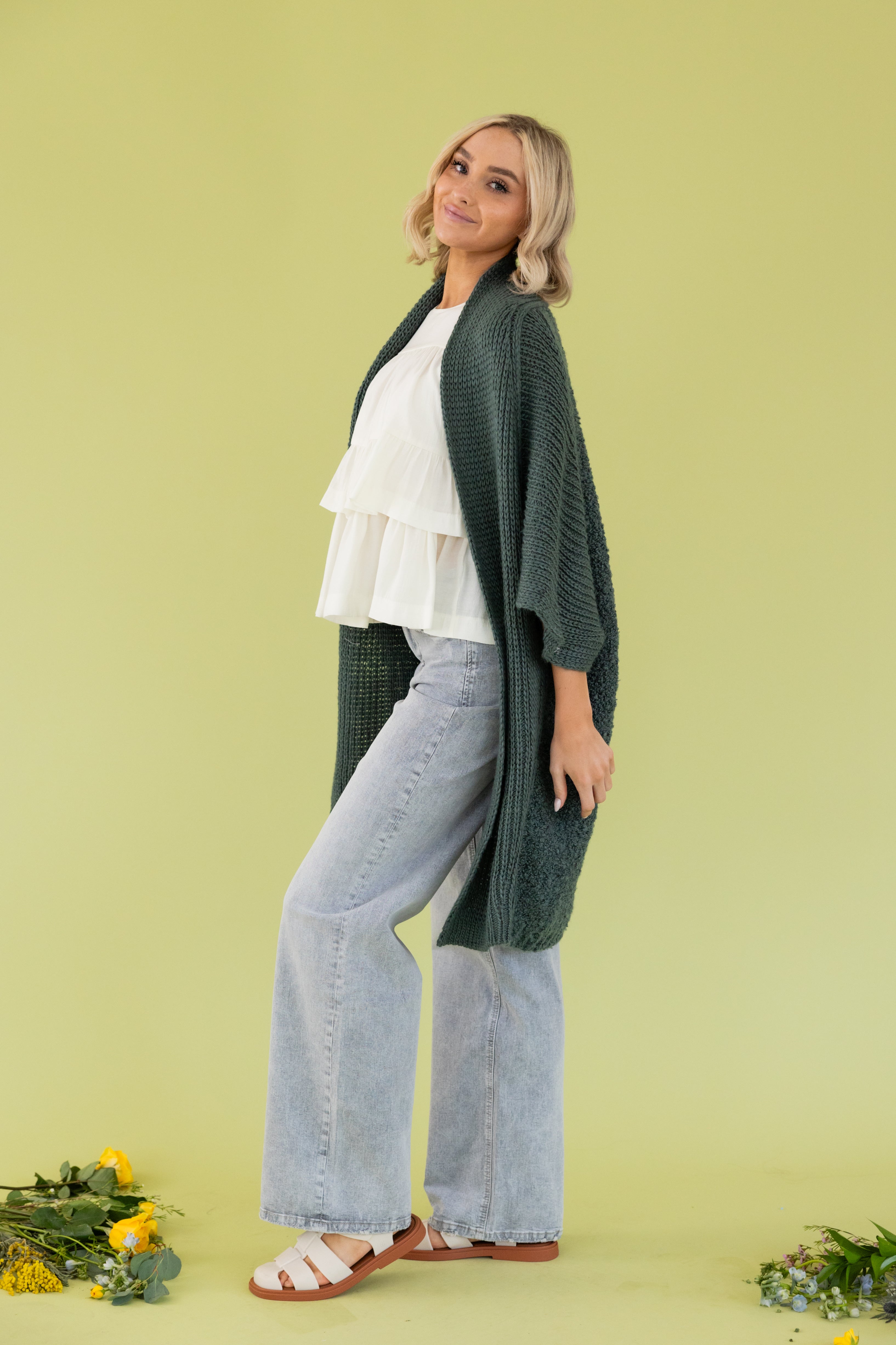 Model is wearing green cardigan in white blouse and grey jeans. 