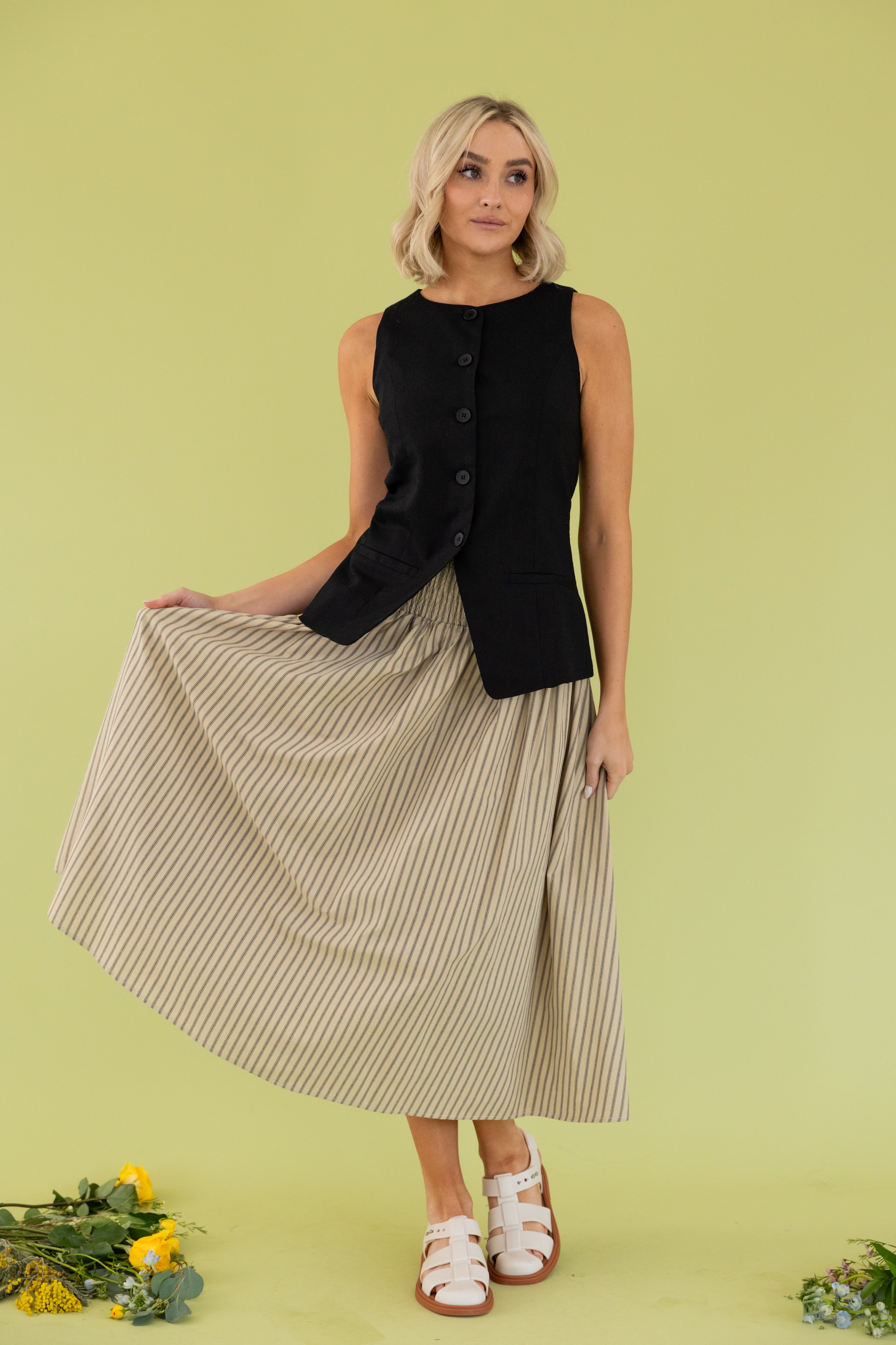 Model is wearing black vest with smocked tan striped midi skirt.