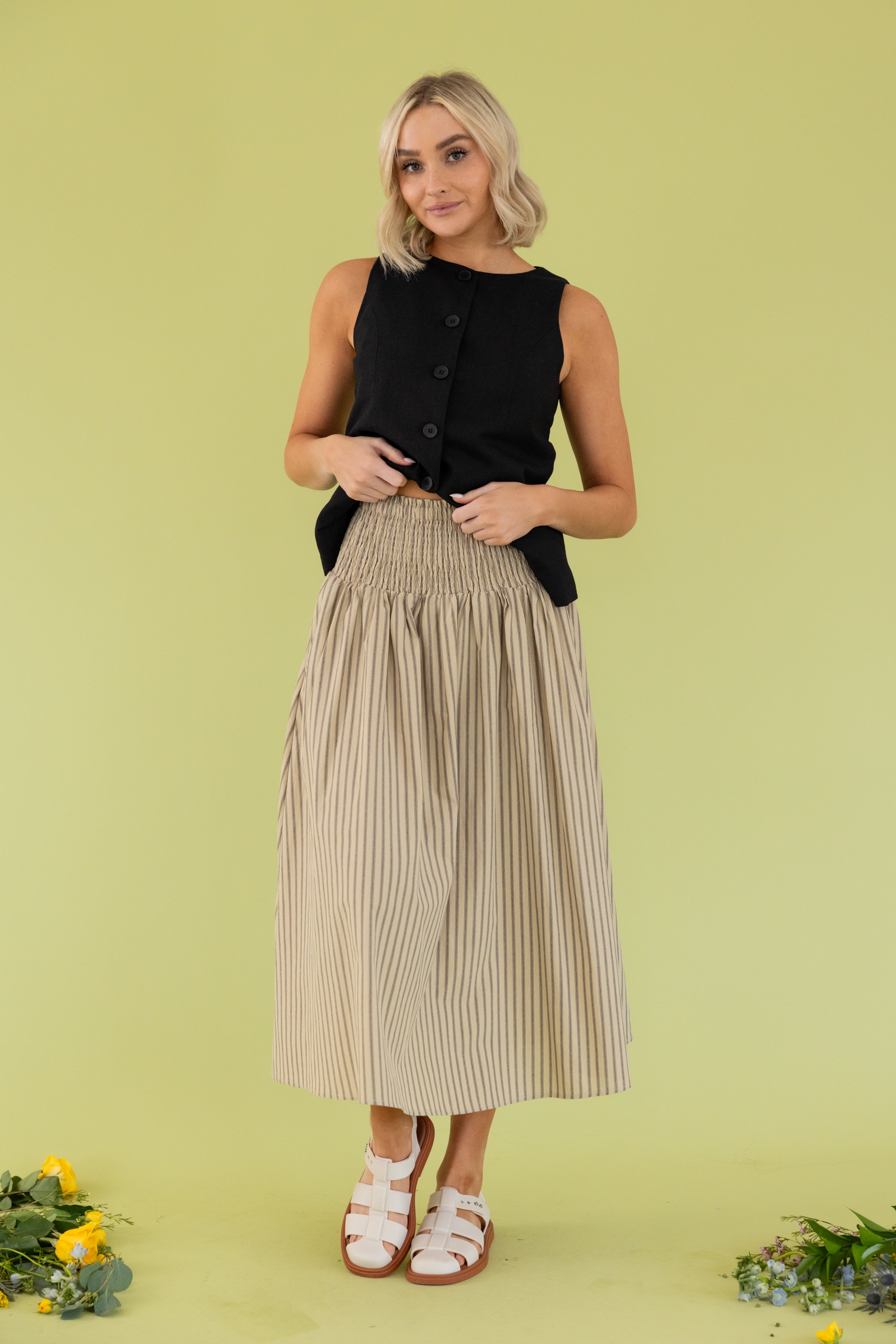 Model is wearing black vest with smocked tan striped midi skirt.